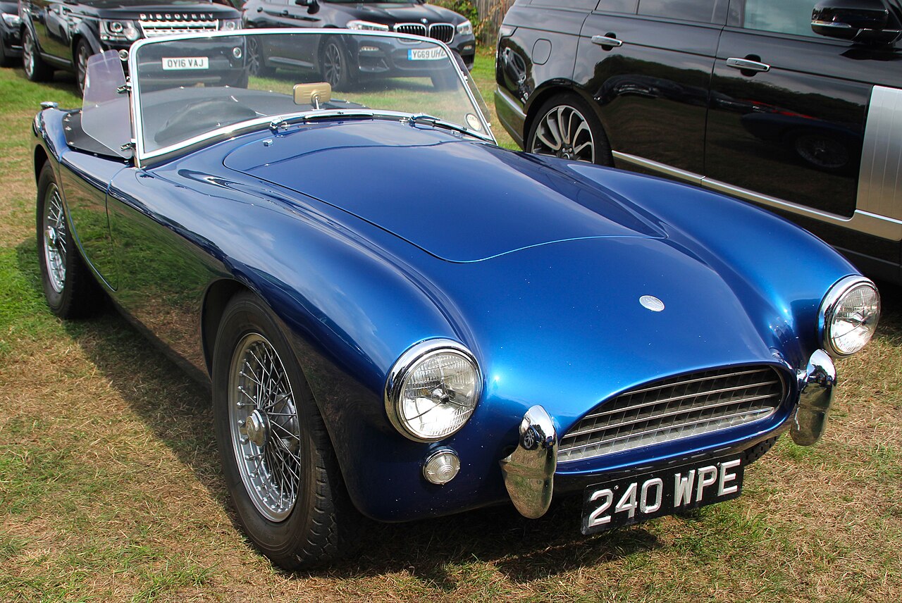 1961 AC Ace in metallic blue with wire wheels and its roof down, front right three-quarter view on grass at a classic car show.