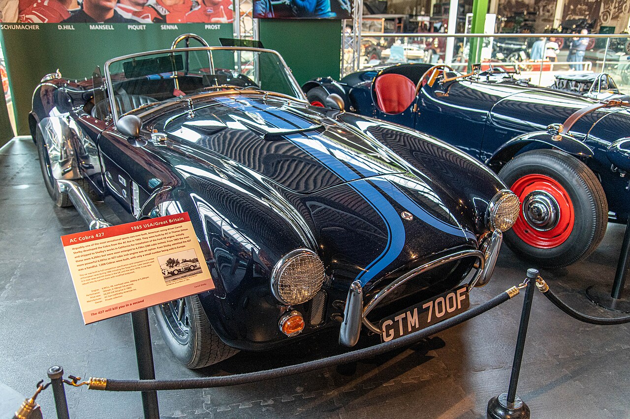 Dark blue AC Cobra 427 with light blue racing stripes, front three-quarter view behind a rope barrier in a museum.
