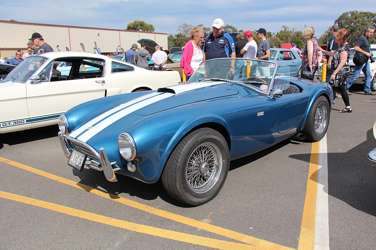 Blue 1963 AC Cobra Mk II 289 with white stripes and wire wheels, roof down, front three-quarter view at a car show.