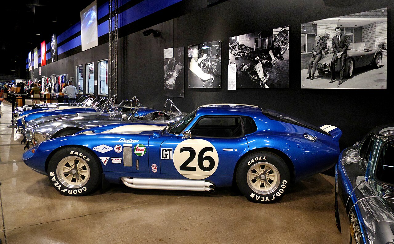 Blue Shelby Cobra Daytona Coupe with race number 26, side-on in a museum with a row of other Cobras behind it.