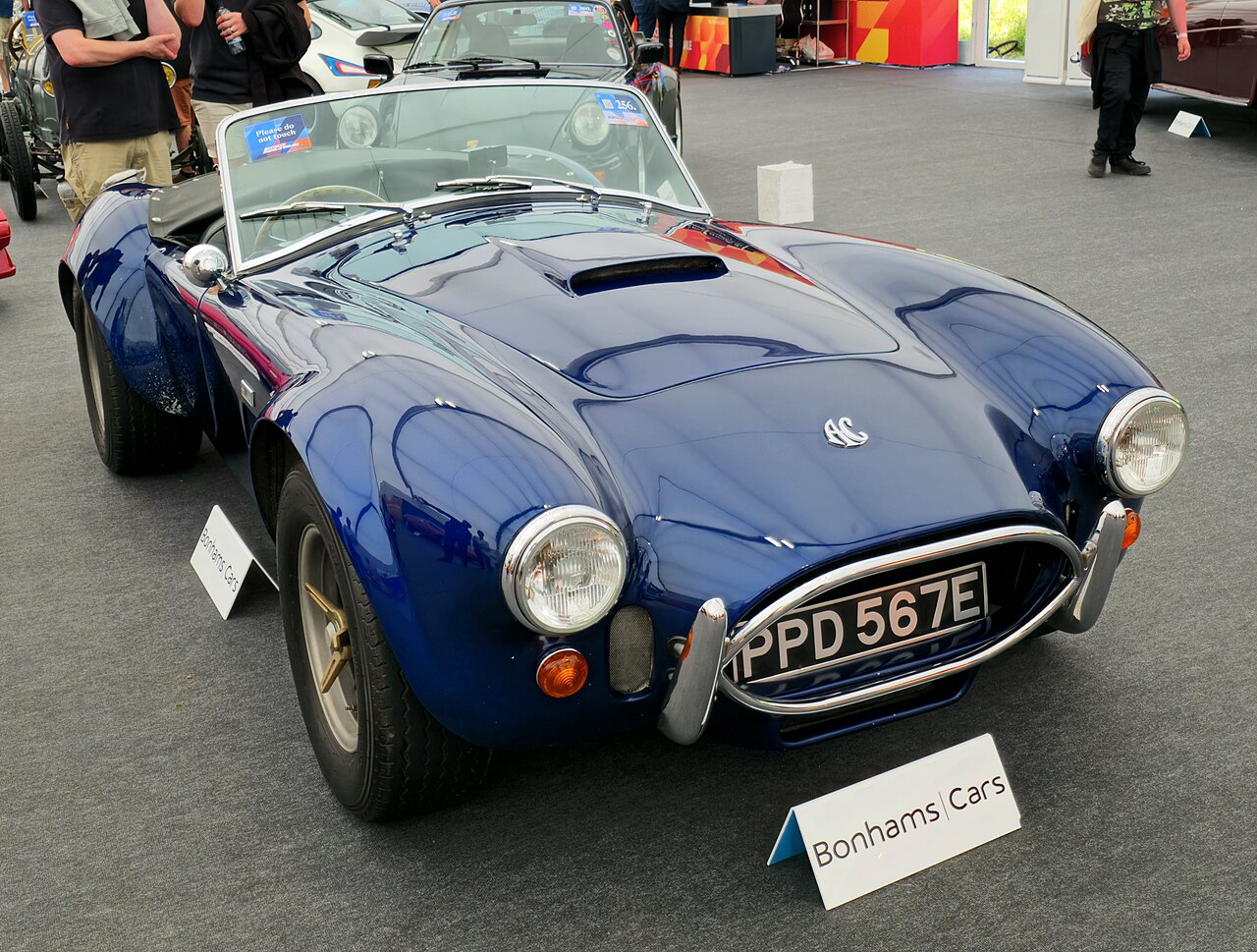 Dark blue 1967 AC Cobra roadster with a bonnet scoop and chrome oval grille, front right three-quarter view on a carpeted show floor.