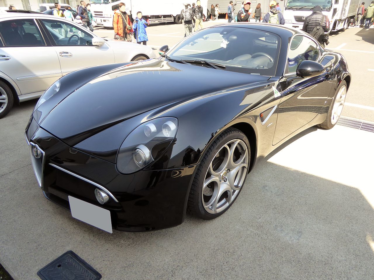 Black Alfa Romeo 8C Competizione coupe, front three-quarter view on a paved lot with visitors in the background.