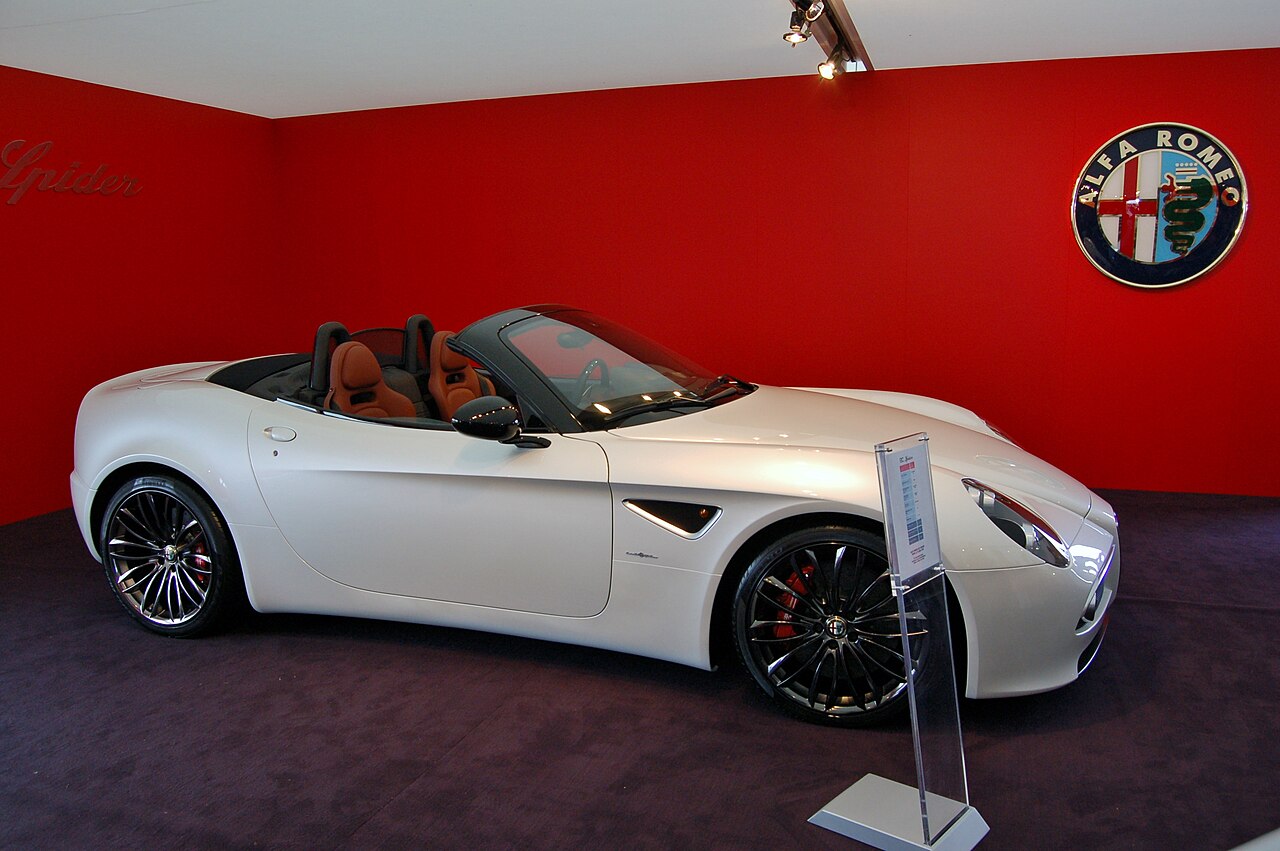 White 2009 Alfa Romeo 8C Spider with its roof down and tan seats, seen from the front right on a red-walled display stand.