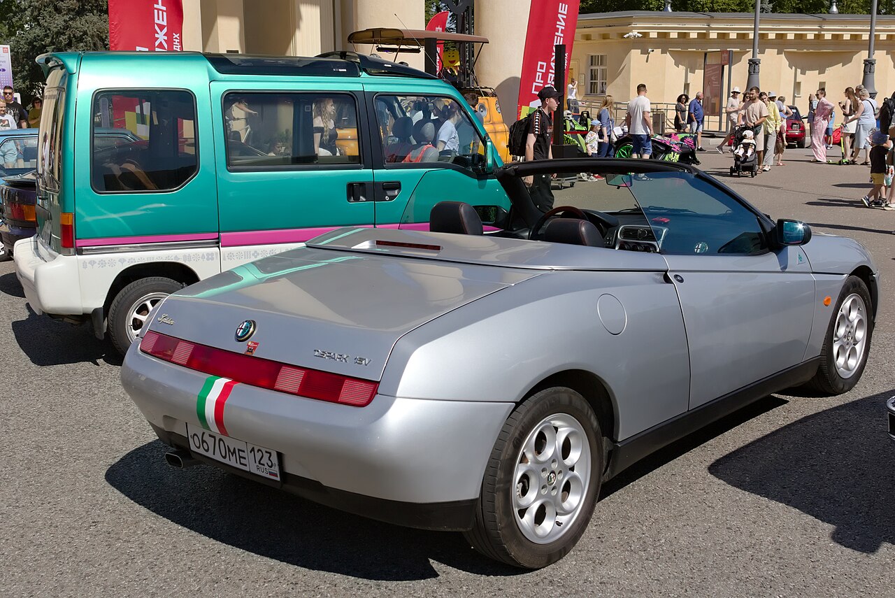Silver Alfa Romeo Spider Type 916 with its roof down, rear right three-quarter view at a crowded outdoor car show.