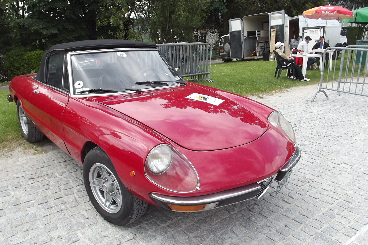 Red Alfa Romeo Spider Duetto with its black soft top up, front right three-quarter view on cobbles at a classic car event.