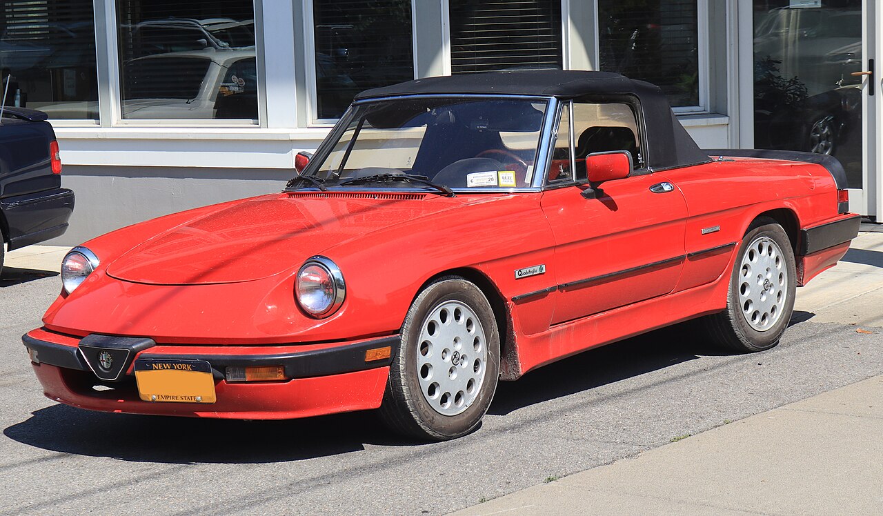 Red 1987 Alfa Romeo Spider Quadrifoglio with its soft top up, front left three-quarter view on a street.