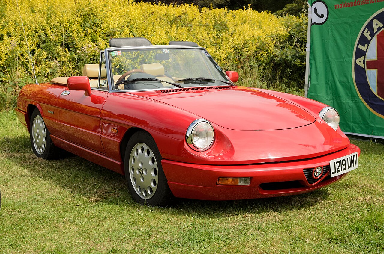 Red 1991 Alfa Romeo Spider Series 4 with its roof down and tan seats, front right three-quarter view on grass at a car show.