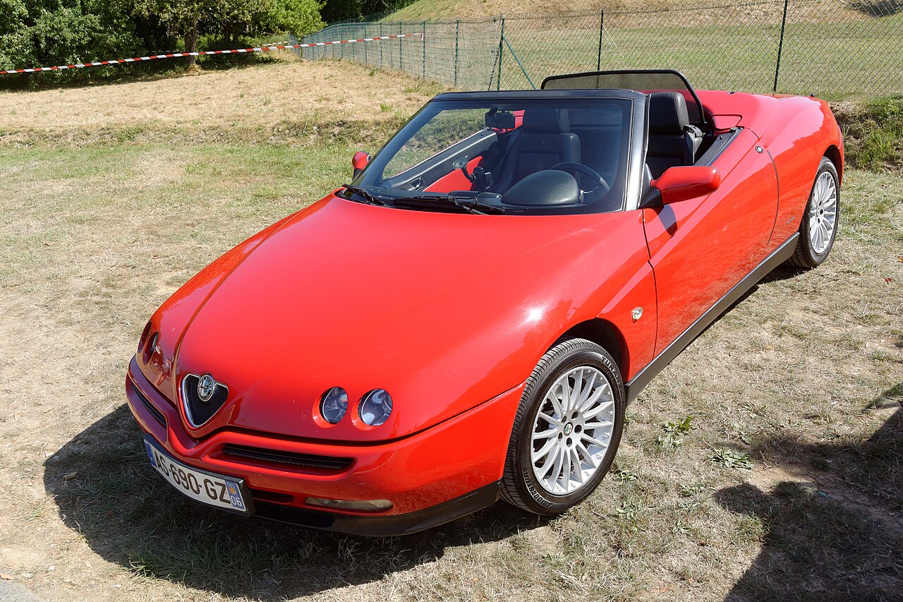 Red Alfa Romeo 916 Spider with its roof down, front left three-quarter view from a high angle on dry grass.