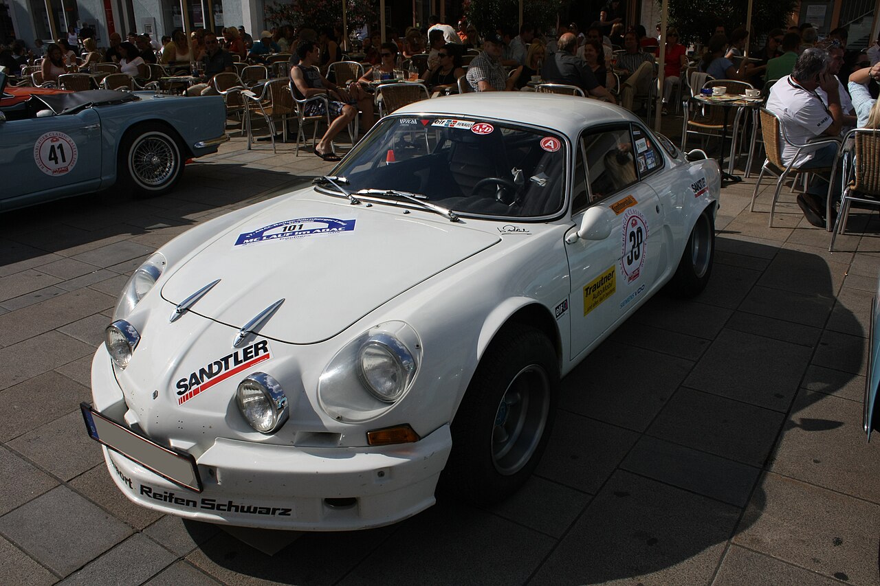 White Alpine A110 rally car with sponsor decals and race numbers, front three-quarter view on a paved square beside a cafe terrace.