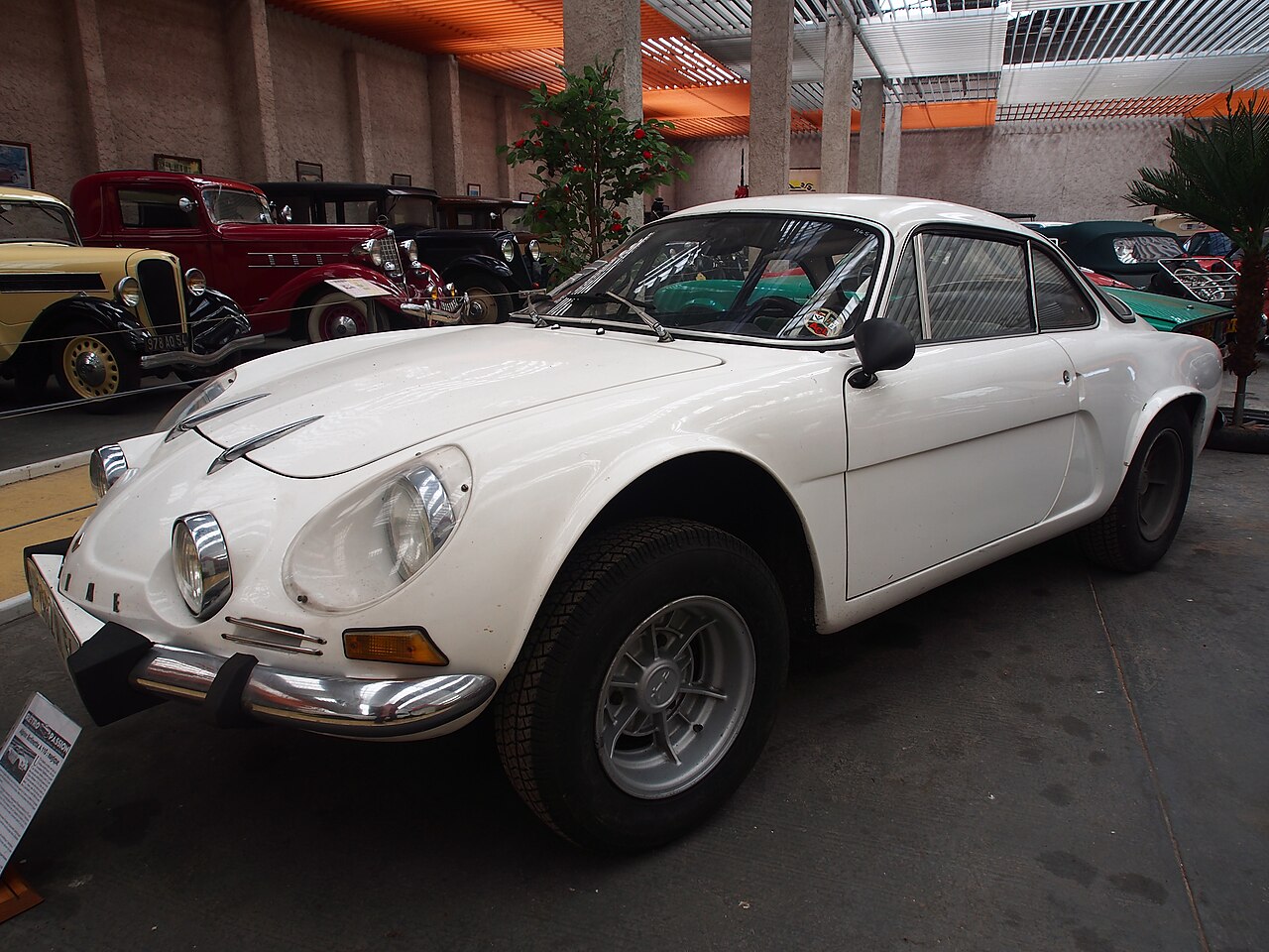White Alpine A110 Berlinette in a museum, front left three-quarter view with older cars parked behind it.