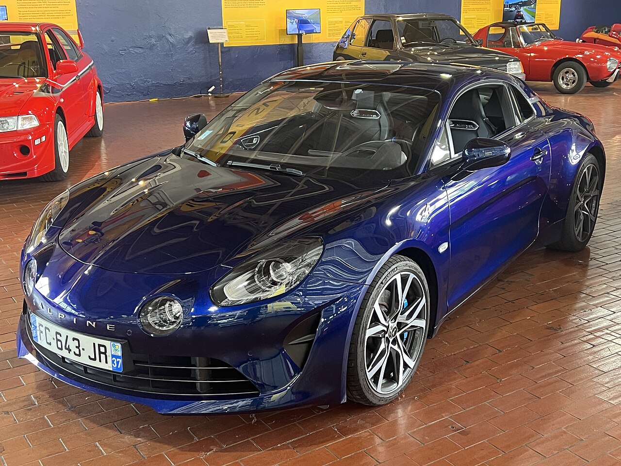 Blue 2018 Alpine A110 coupe, front three-quarter view from a raised angle on a brick floor in the Lane Motor Museum.