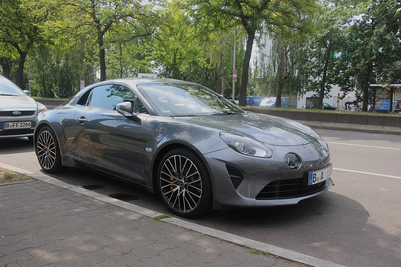 Grey Alpine A110 GT with multi-spoke alloy wheels, front three-quarter view parked at a kerb on a tree-lined street.