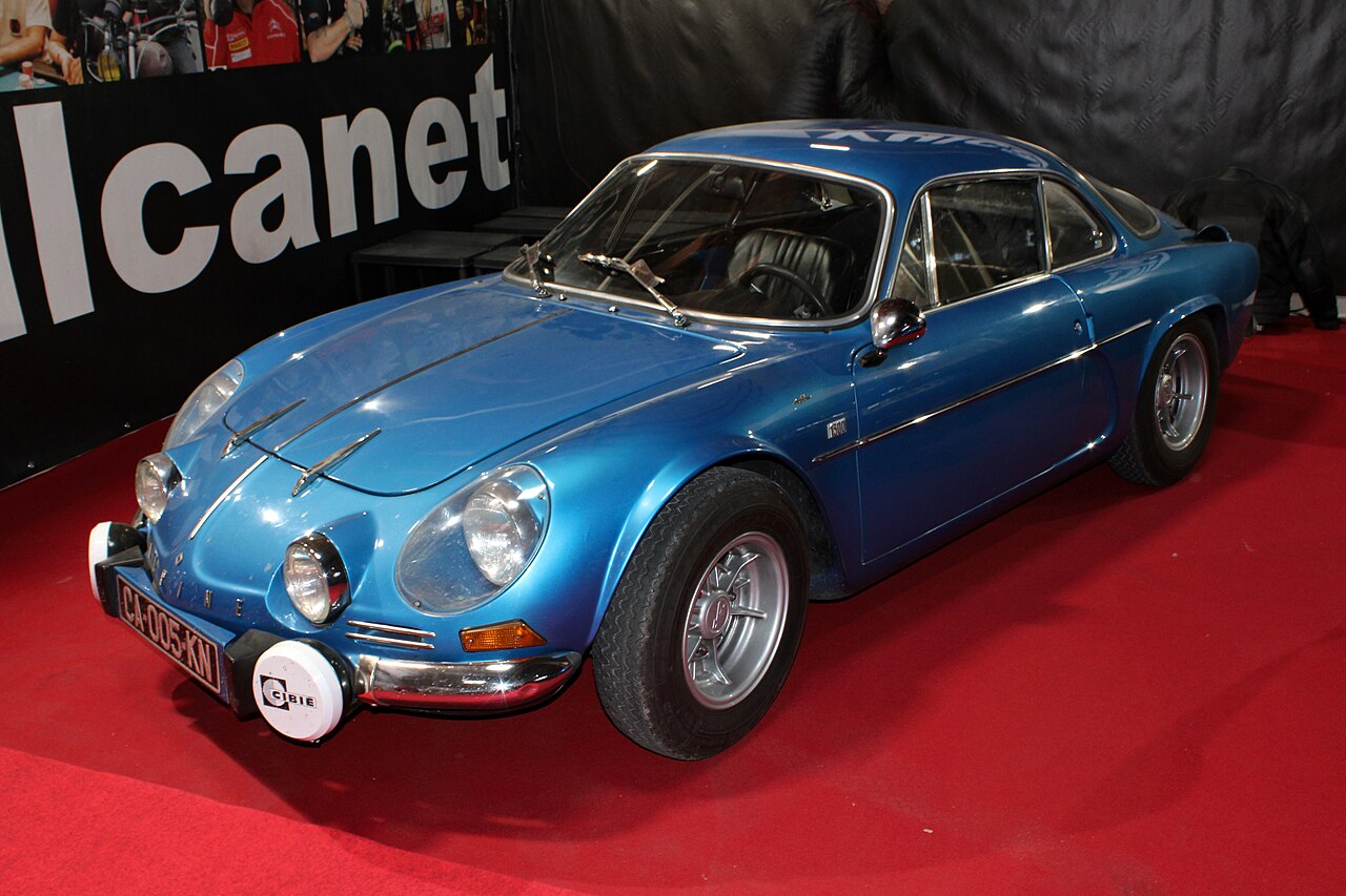 Blue classic Alpine A110 Berlinette with a white driving-lamp cover, front three-quarter view on a red carpet at a motor show.