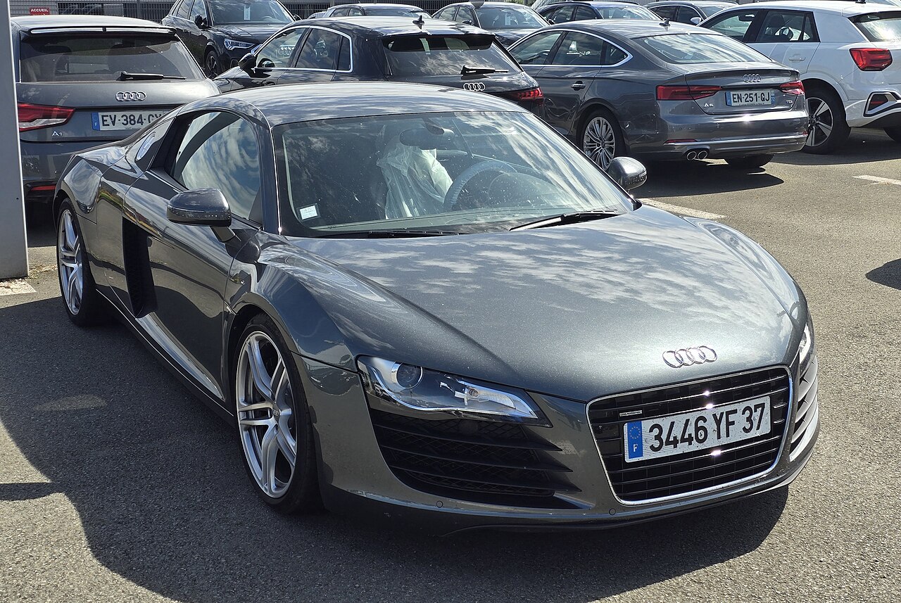 Grey Audi R8 coupe, front right three-quarter view in a car park with other Audi cars behind.