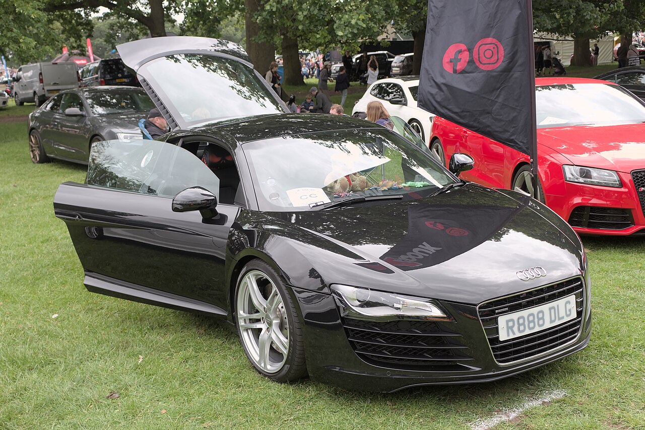 Black Audi R8 coupe with its door and rear engine cover open, front three-quarter view on grass at a car show.