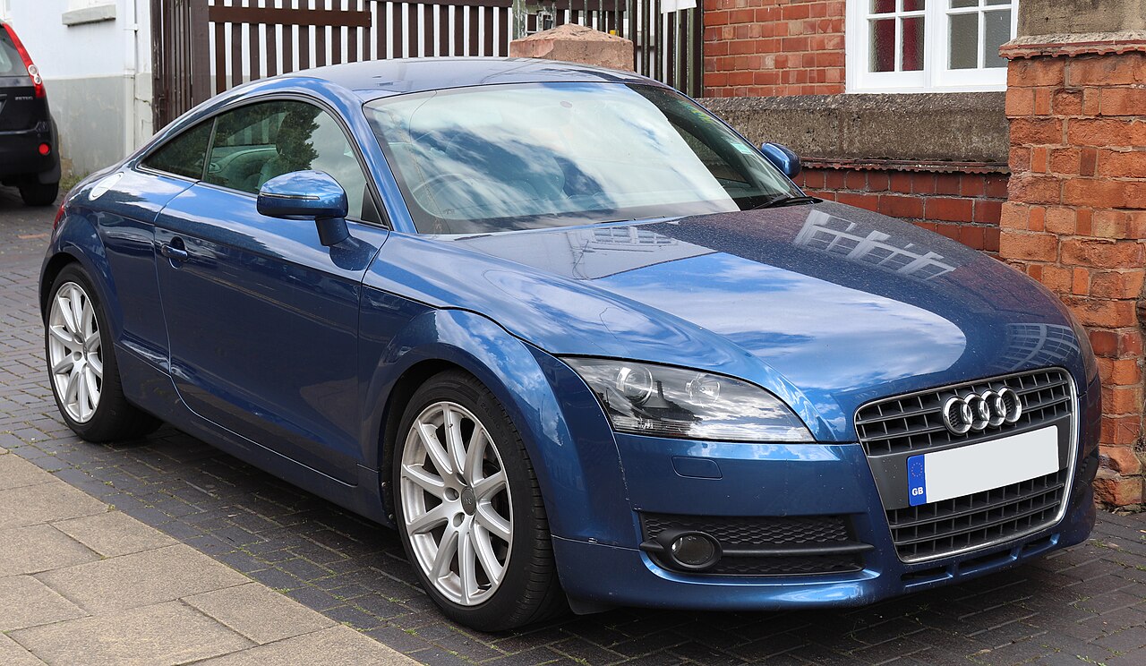 Blue 2008 Audi TT coupe, front three-quarter view parked on a brick-paved street beside a red brick wall.