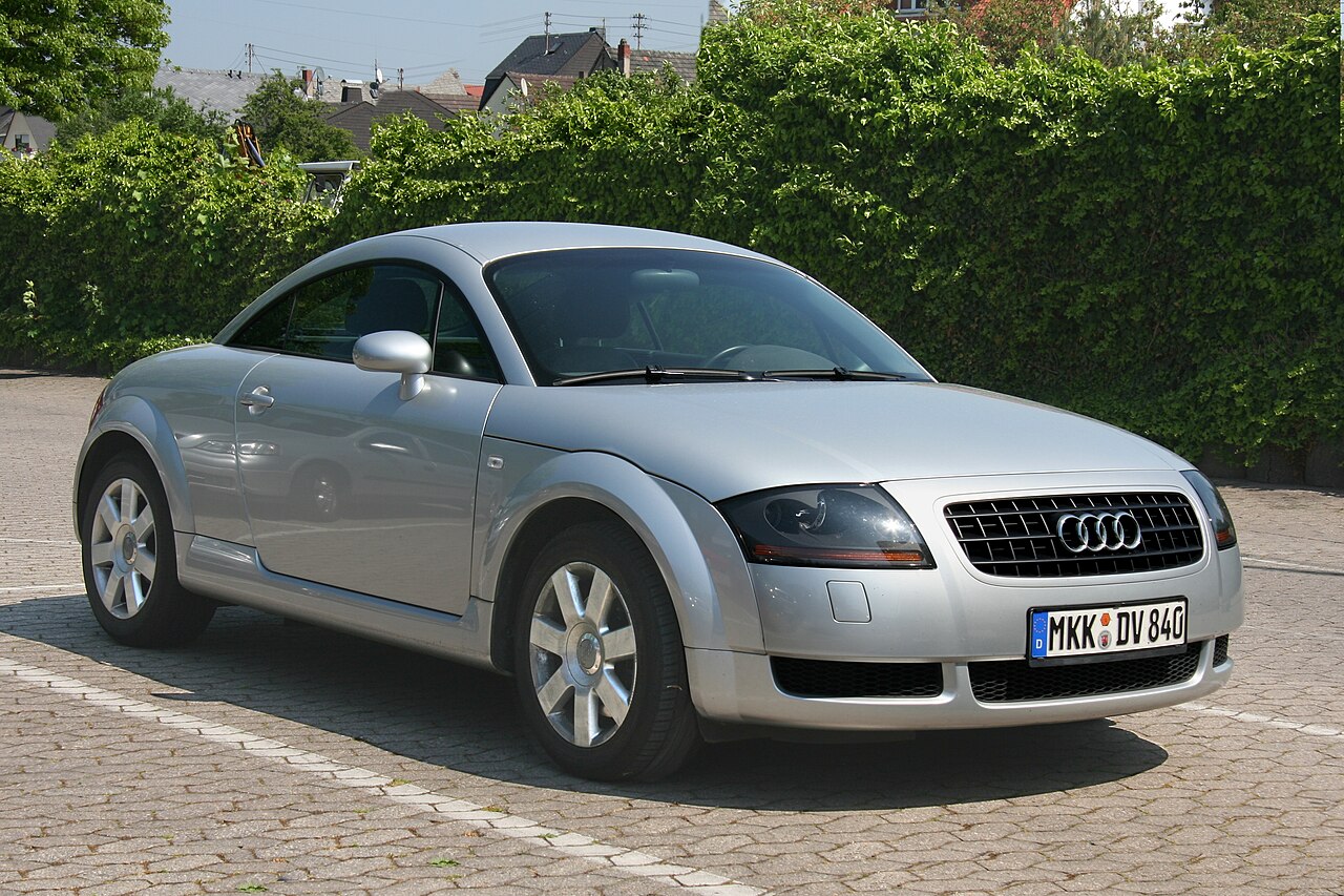 Silver first-generation Audi TT coupe, front three-quarter view on a paved car park beside a hedge.