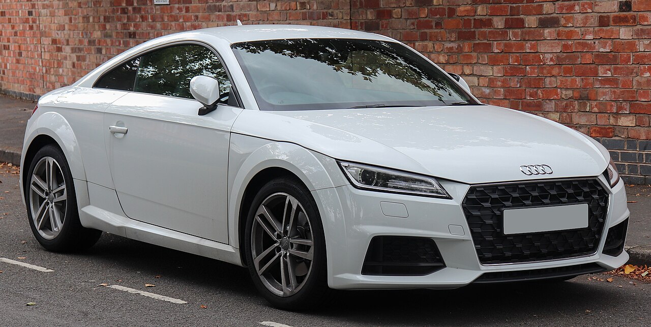 White 2019 Audi TT Sport coupe, front three-quarter view parked on a street beside a red brick wall.