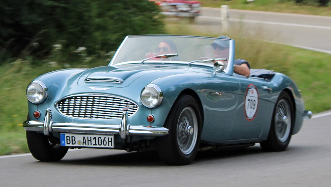 Pale blue 1957 Austin-Healey 100-6 with its roof down, front left three-quarter view as it is driven along a road.