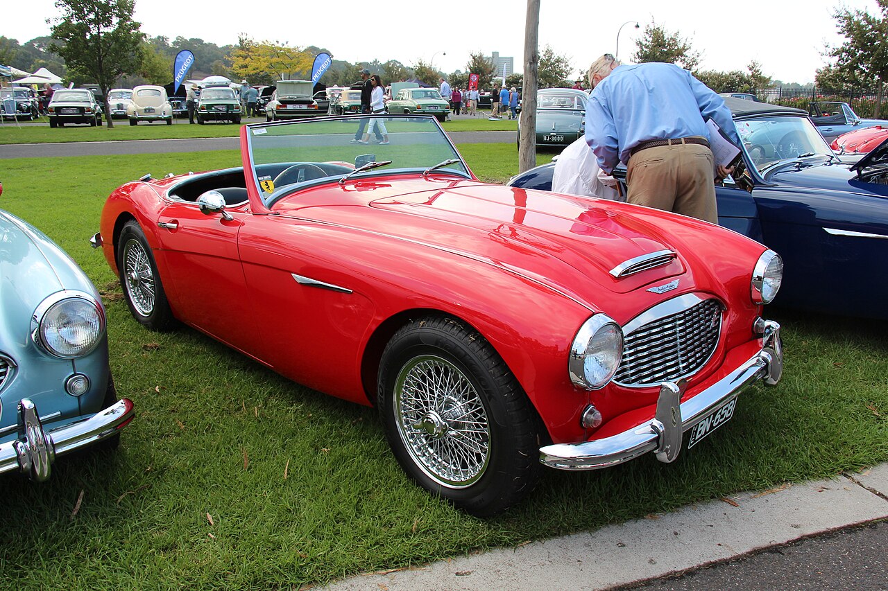 Red 1958 Austin-Healey 100-6 with its roof down and wire wheels, front three-quarter view on grass at a car show.