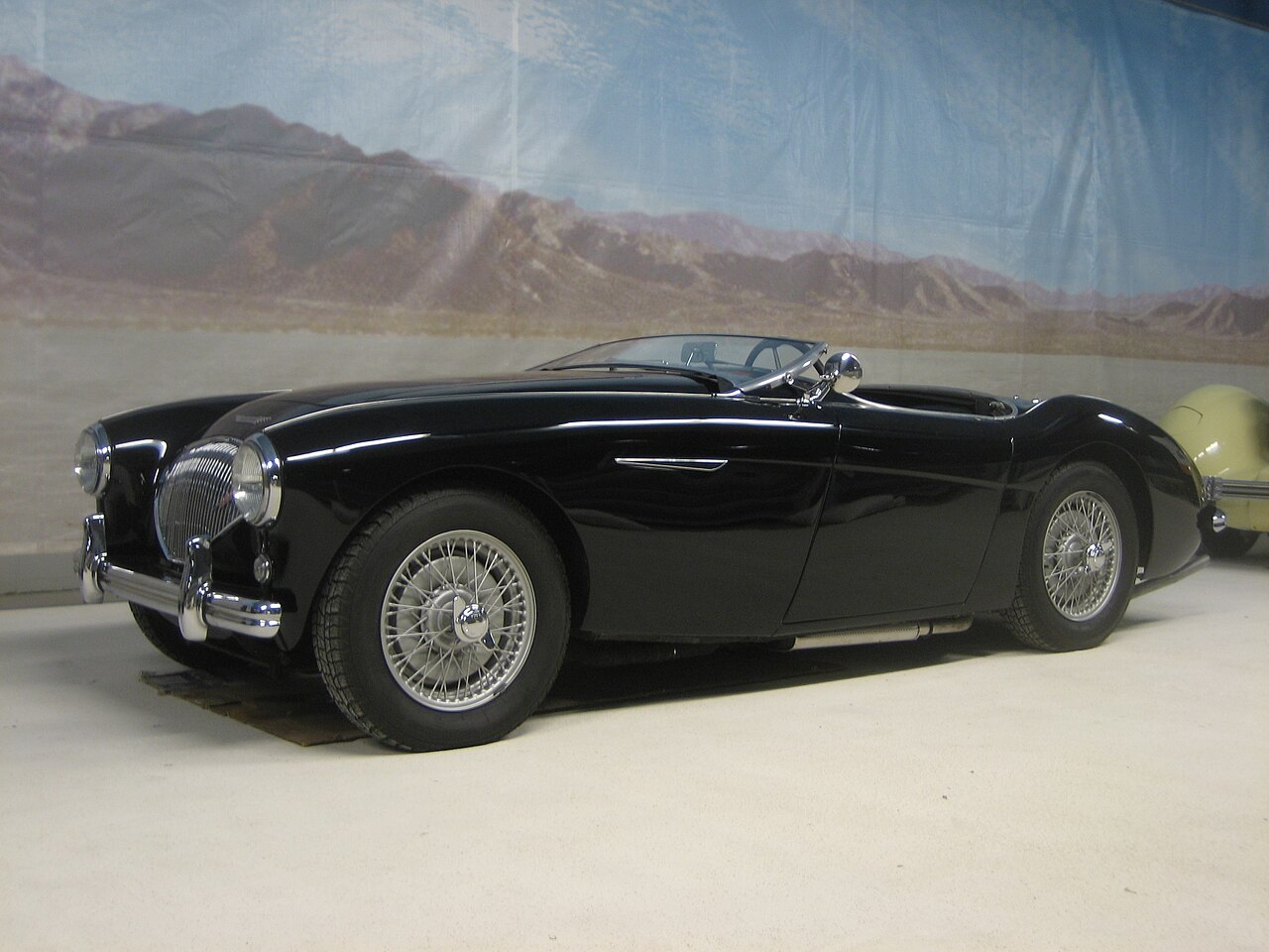 Black 1954 Austin-Healey 100 BN1 with wire wheels and its roof down, front left three-quarter view in a museum.