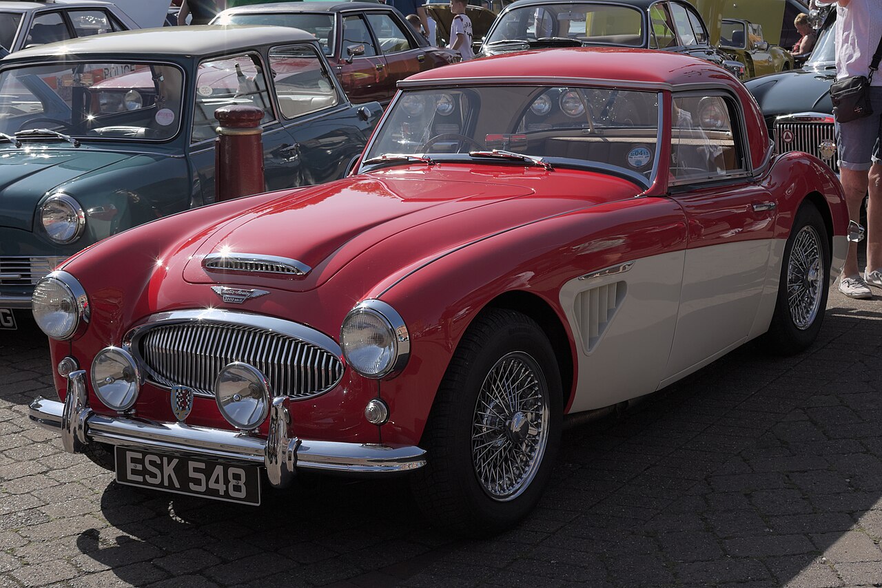 Red and white Austin-Healey 3000 Mk II with a hardtop and wire wheels, front three-quarter view at a car show.