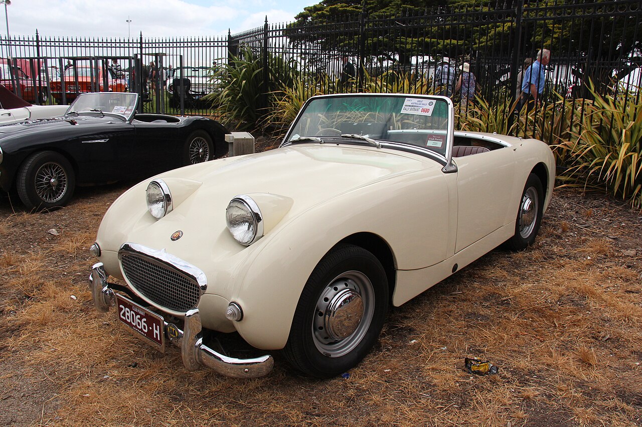 Cream 1959 Austin-Healey Sprite Mk I, the Frogeye, with its roof down, front three-quarter view on dry grass by an iron fence.
