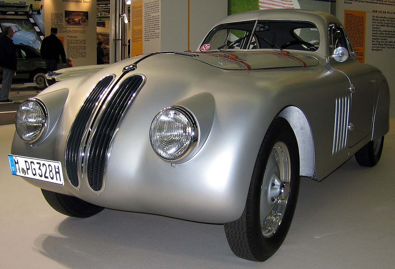Silver BMW 328 Touring Coupe, a streamlined Mille Miglia racing car, in front three-quarter view on an indoor exhibition stand.