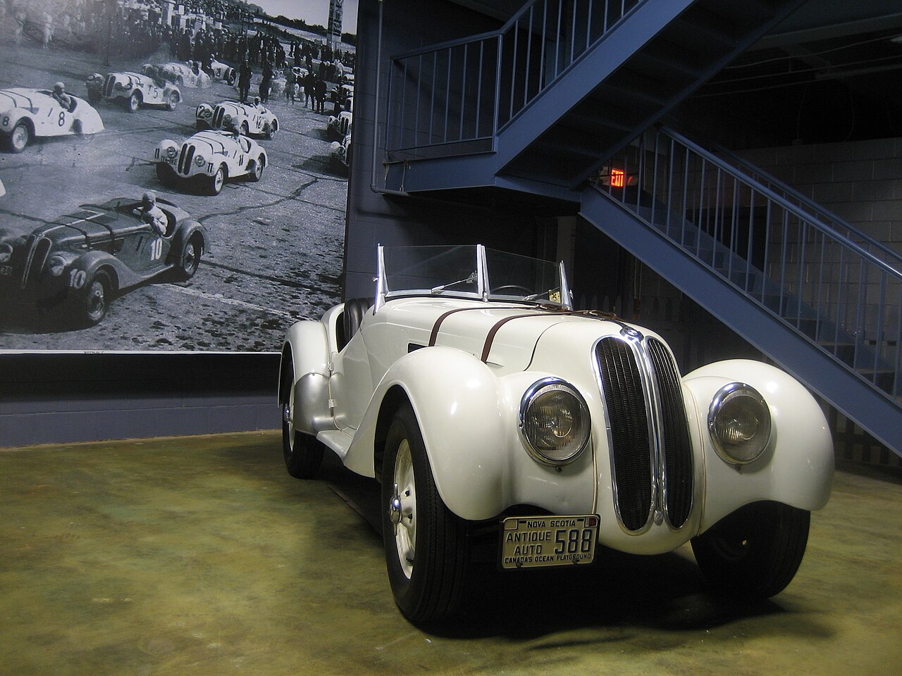 Cream-white 1939 BMW 328 roadster, front right three-quarter view in a museum with a large black-and-white racing photograph behind it.