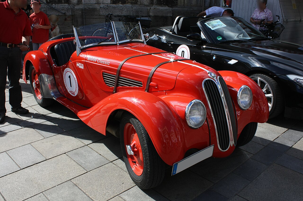 Red BMW 328 roadster wearing rally numbers, front three-quarter view on paving stones beside a black BMW roadster.