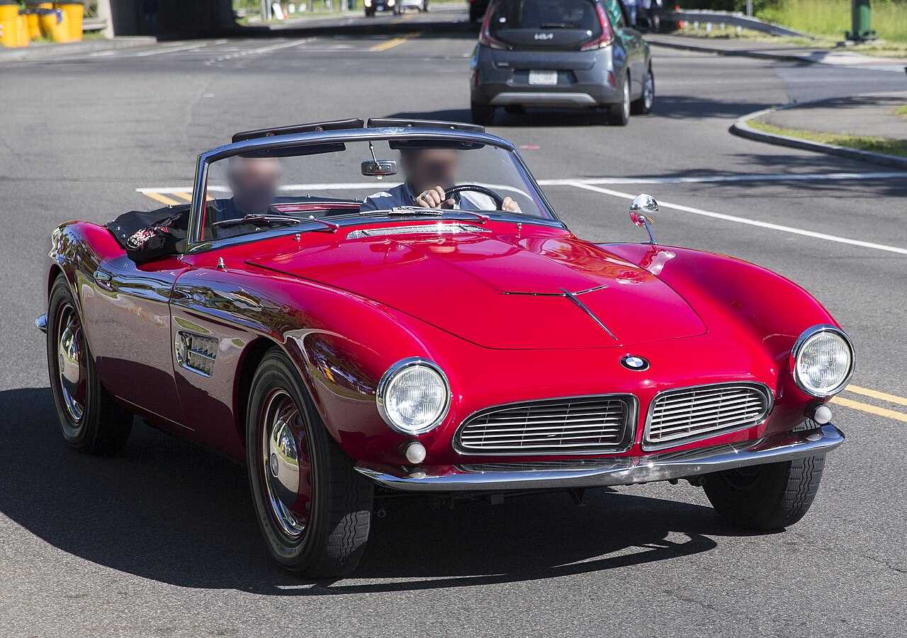 Red 1957 BMW 507 roadster with its roof down, front three-quarter view driving along a street.