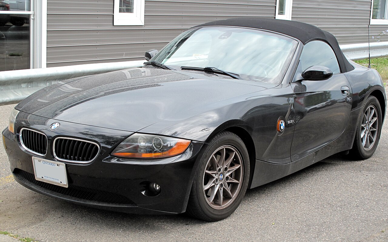 2004 BMW Z4 in black with its soft top up, front three-quarter view beside a guardrail.