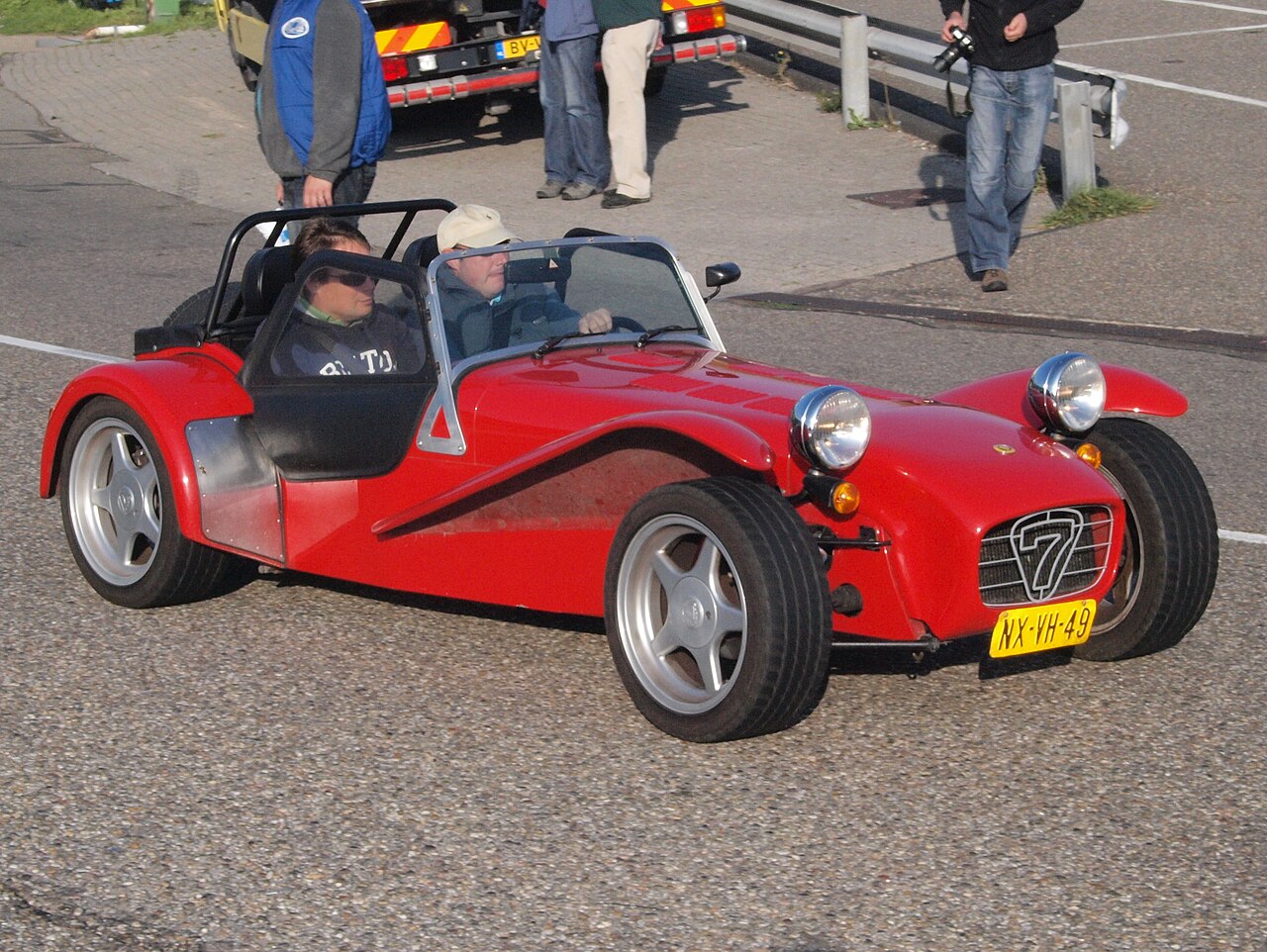 Red Caterham Super Seven HPC with silver alloy wheels and two occupants, front right three-quarter view on a road.