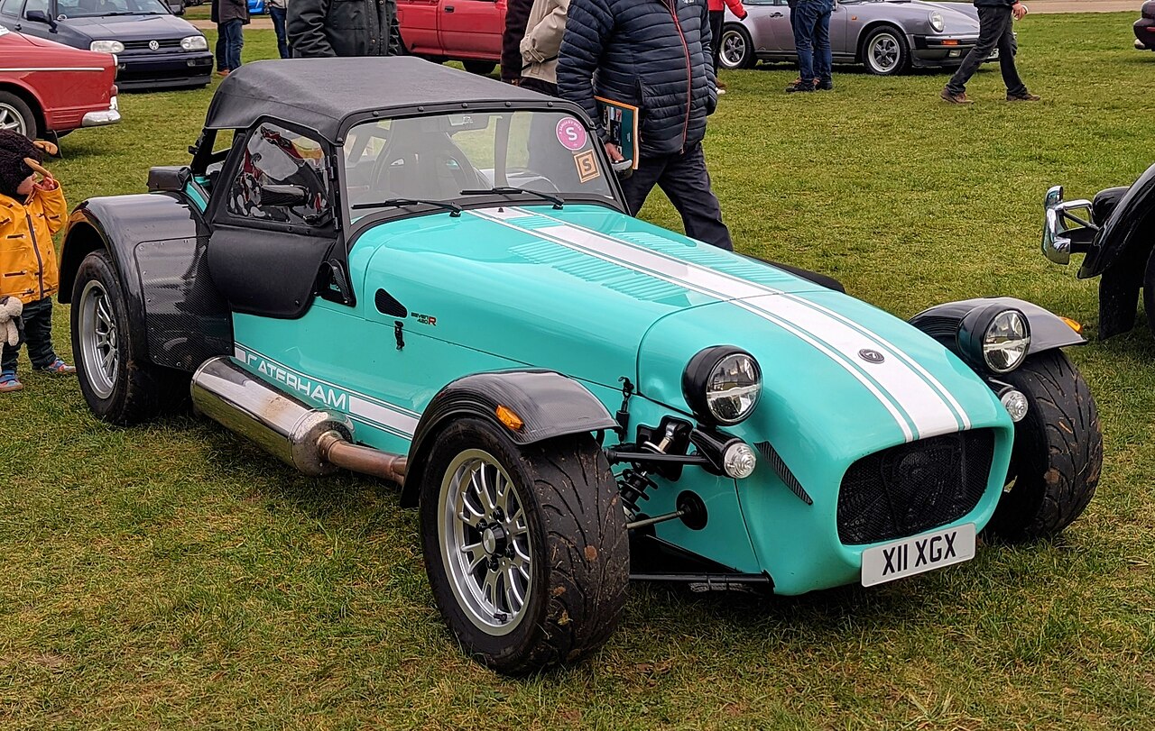 2020 Caterham Seven 420R in turquoise with a white bonnet stripe and its black soft top up, front three-quarter view on grass.