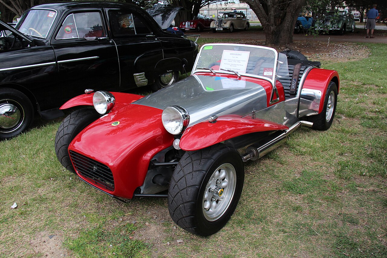 Red 1962 Lotus Super Seven Series 2 with a polished aluminium bonnet and wide tyres, front left three-quarter view on grass at a car show.