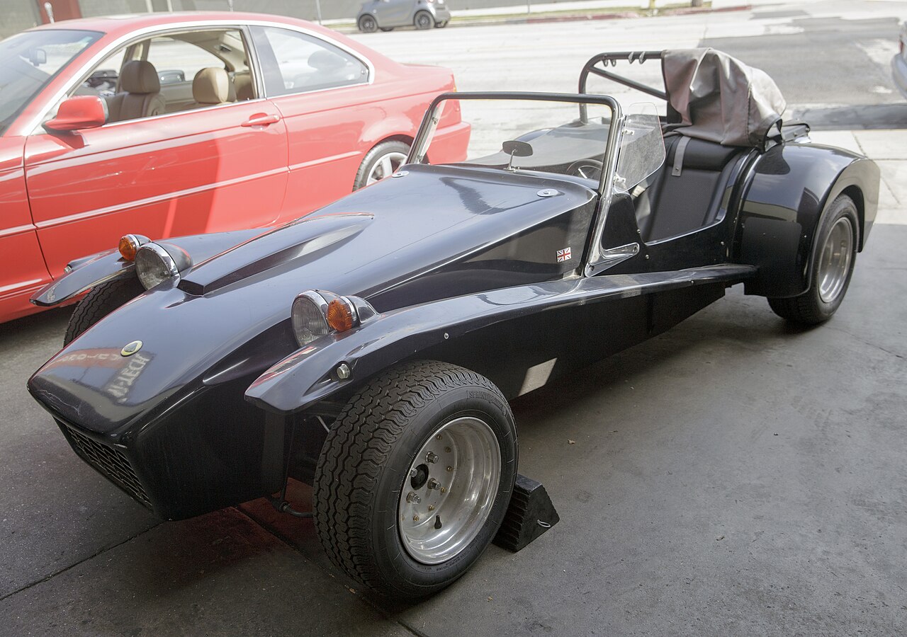 Black Lotus Super Seven S4 roadster, front left three-quarter view parked in a garage bay next to a red coupe.