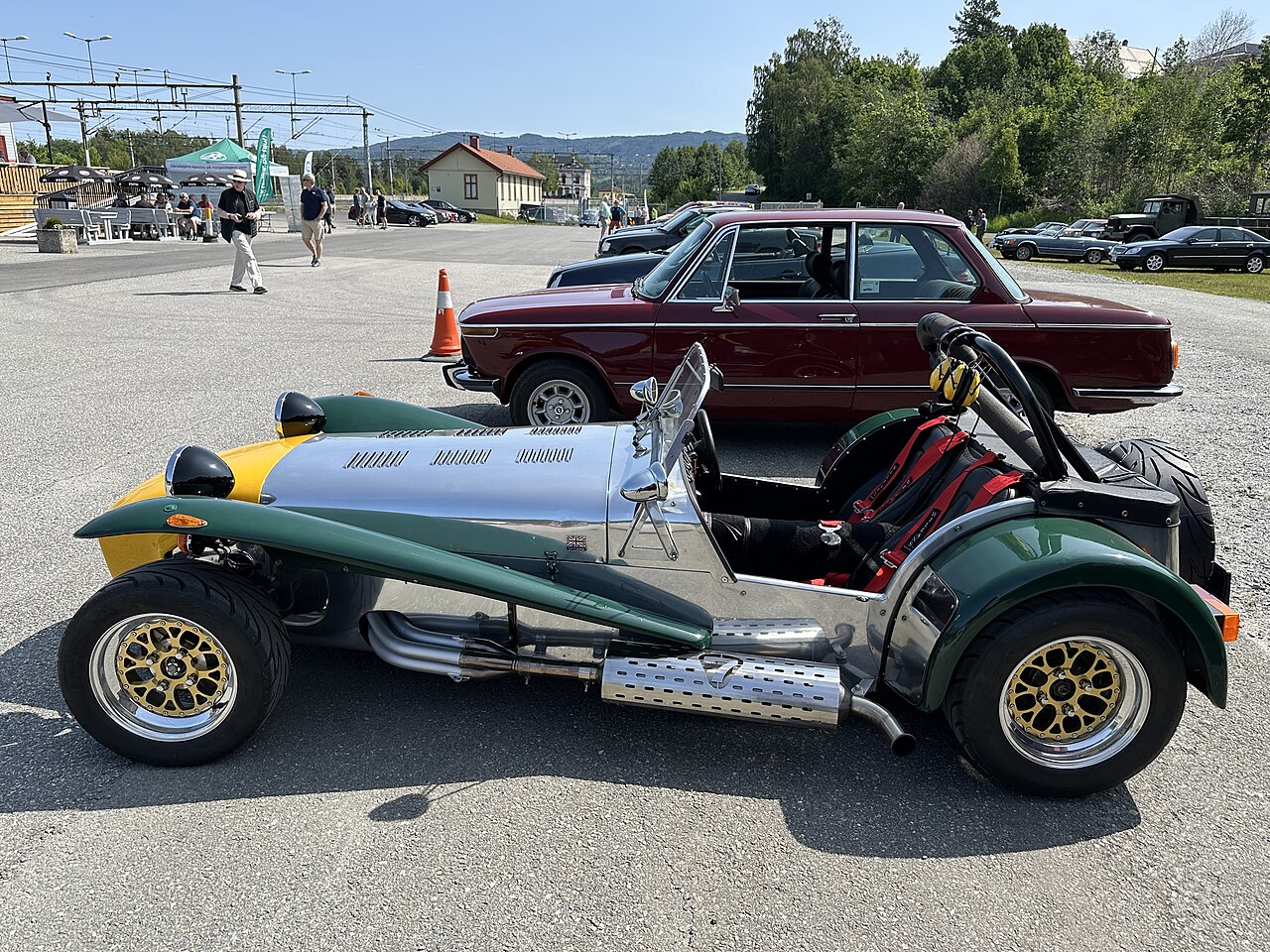 1987 Caterham Seven with polished aluminium panels, a yellow nose and green wings, side-on in a car park.