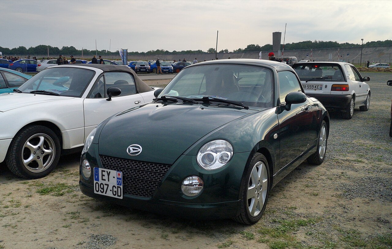 Dark green Daihatsu Copen with its roof up, front left three-quarter view parked on gravel at a car meet.