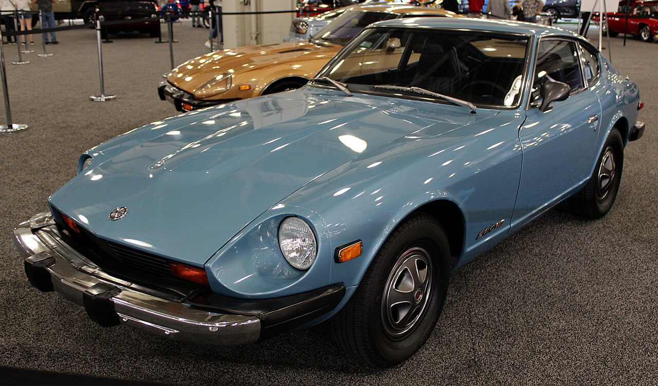 Pale blue 1976 Datsun 280Z with large chrome bumpers, front three-quarter view on carpet at an indoor auto show.