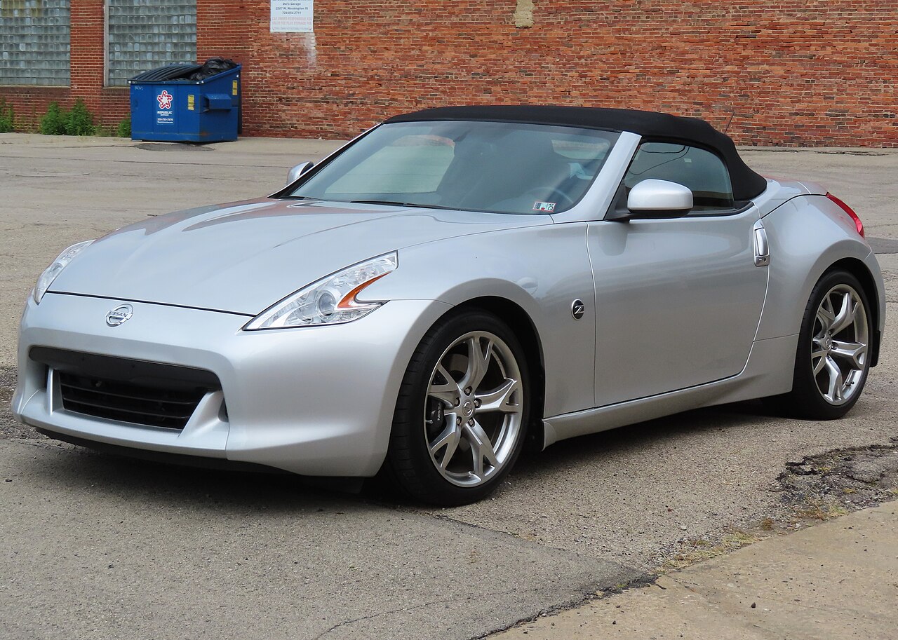 2010 Nissan 370Z Touring roadster in silver with its black soft top up, front left three-quarter view in a car park by a brick wall.