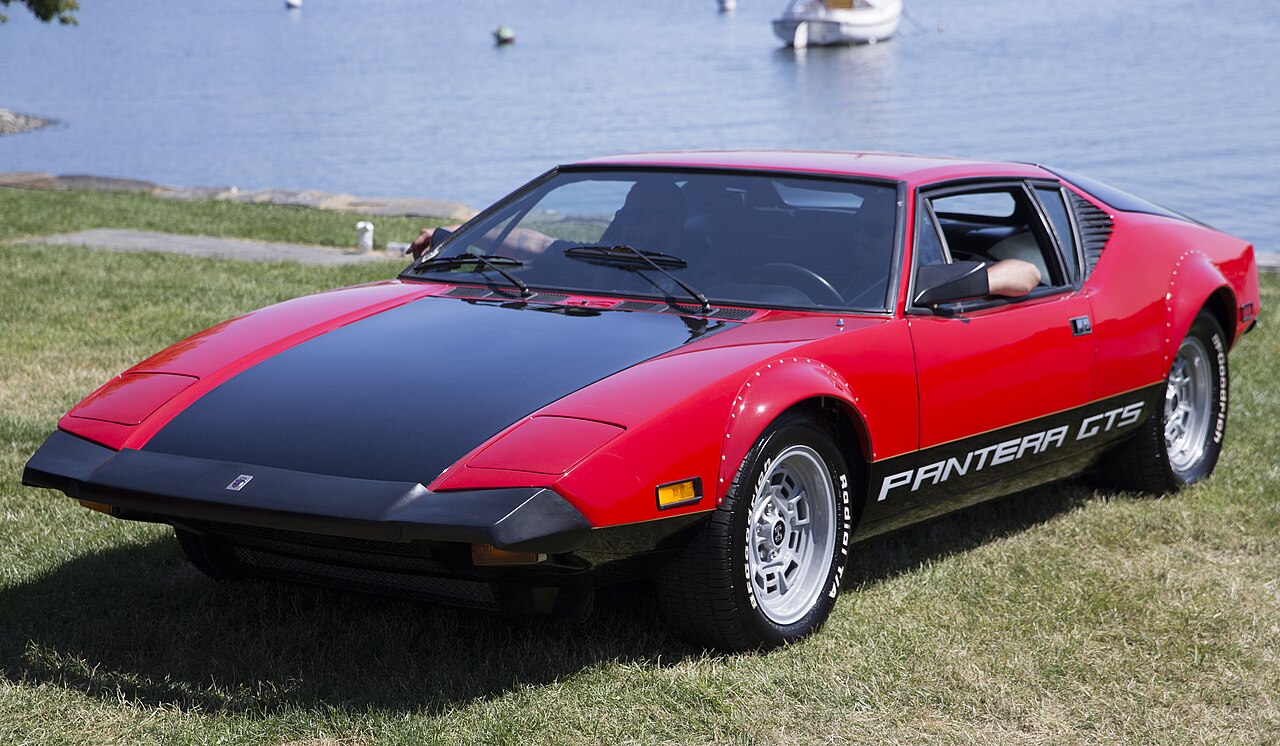 Red 1974 De Tomaso Pantera GTS with a black bonnet and side script, front three-quarter view on grass by the water.