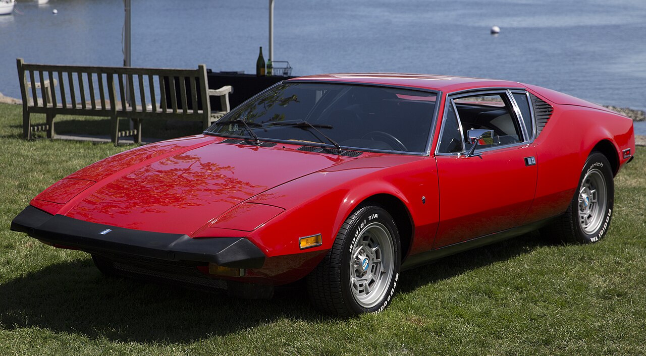 Red 1974 De Tomaso Pantera L, front left three-quarter view on a lawn with a bench and water behind.