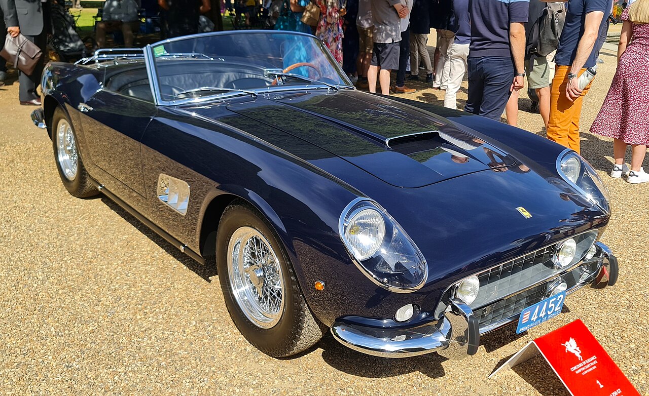 Dark blue 1961 Ferrari 250 GT California Spyder with its roof down and wire wheels, front right three-quarter view on gravel at a concours.