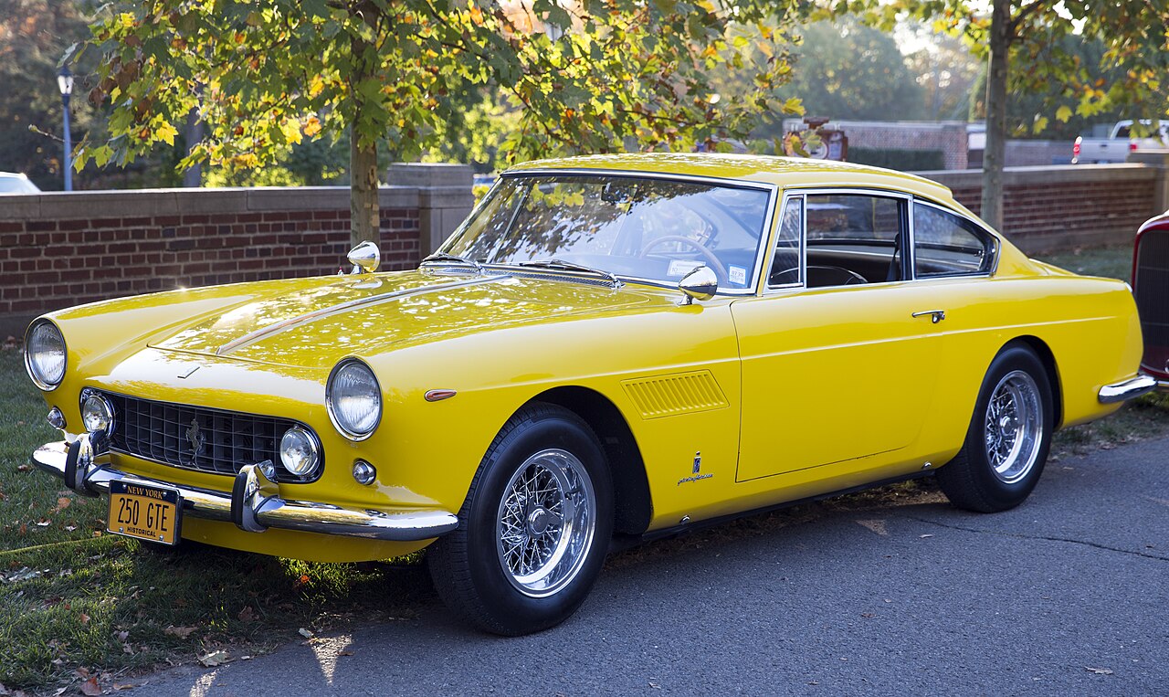 Yellow 1961 Ferrari 250 GTE Series I with wire wheels, seen from the front left parked by a brick wall.