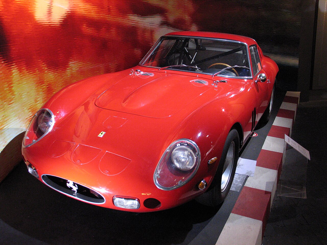 Red Ferrari 250 GTO, front three-quarter view on display indoors against an orange backdrop.