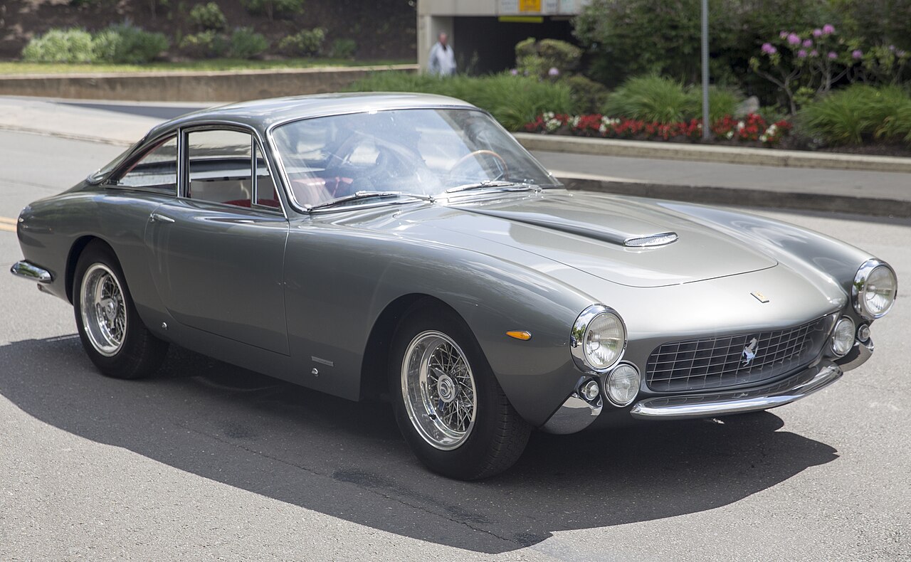 Grey Ferrari 250 GT Lusso with wire wheels, front three-quarter view on a street beside a flower bed.