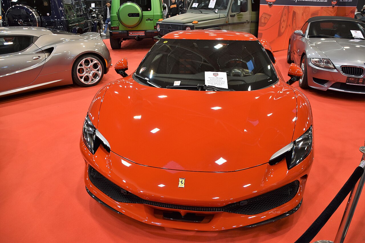 Orange 2022 Ferrari 296 GTB seen head-on on a red carpet at an indoor car show, with other cars parked around it.