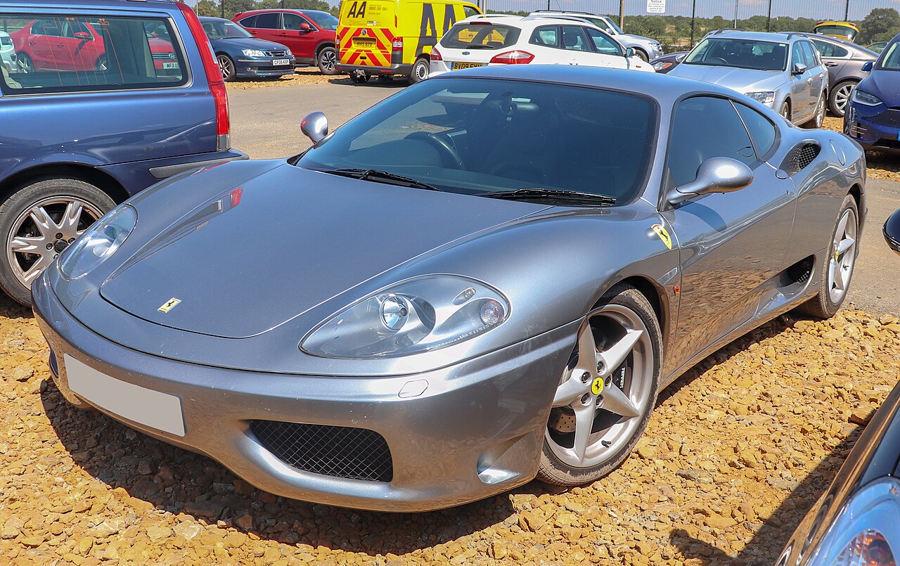Silver-grey 2003 Ferrari 360 Modena, front left three-quarter view on gravel with other cars parked around it.