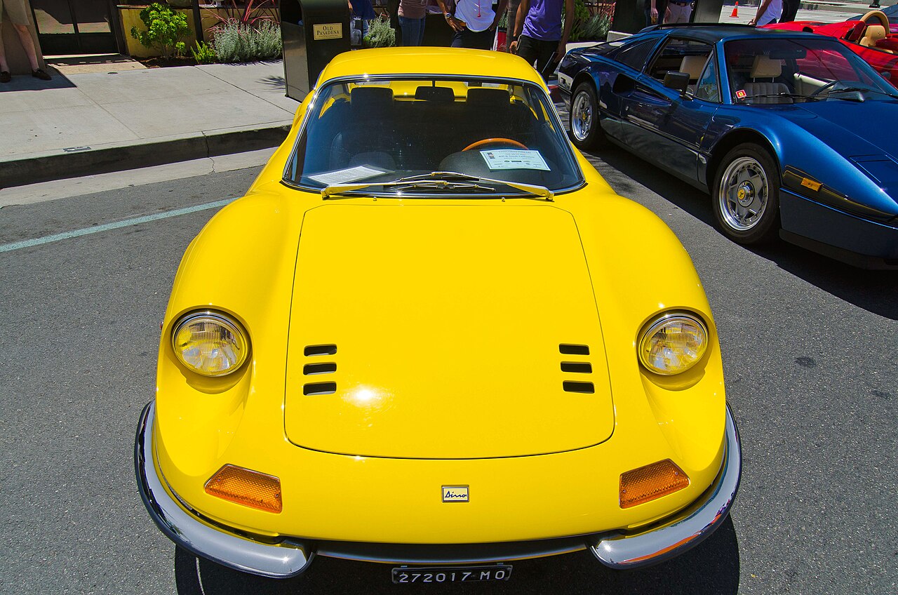 Yellow 1968 Ferrari Dino 206 GT in a head-on view showing its round headlamps and vented bonnet, at a street car show.