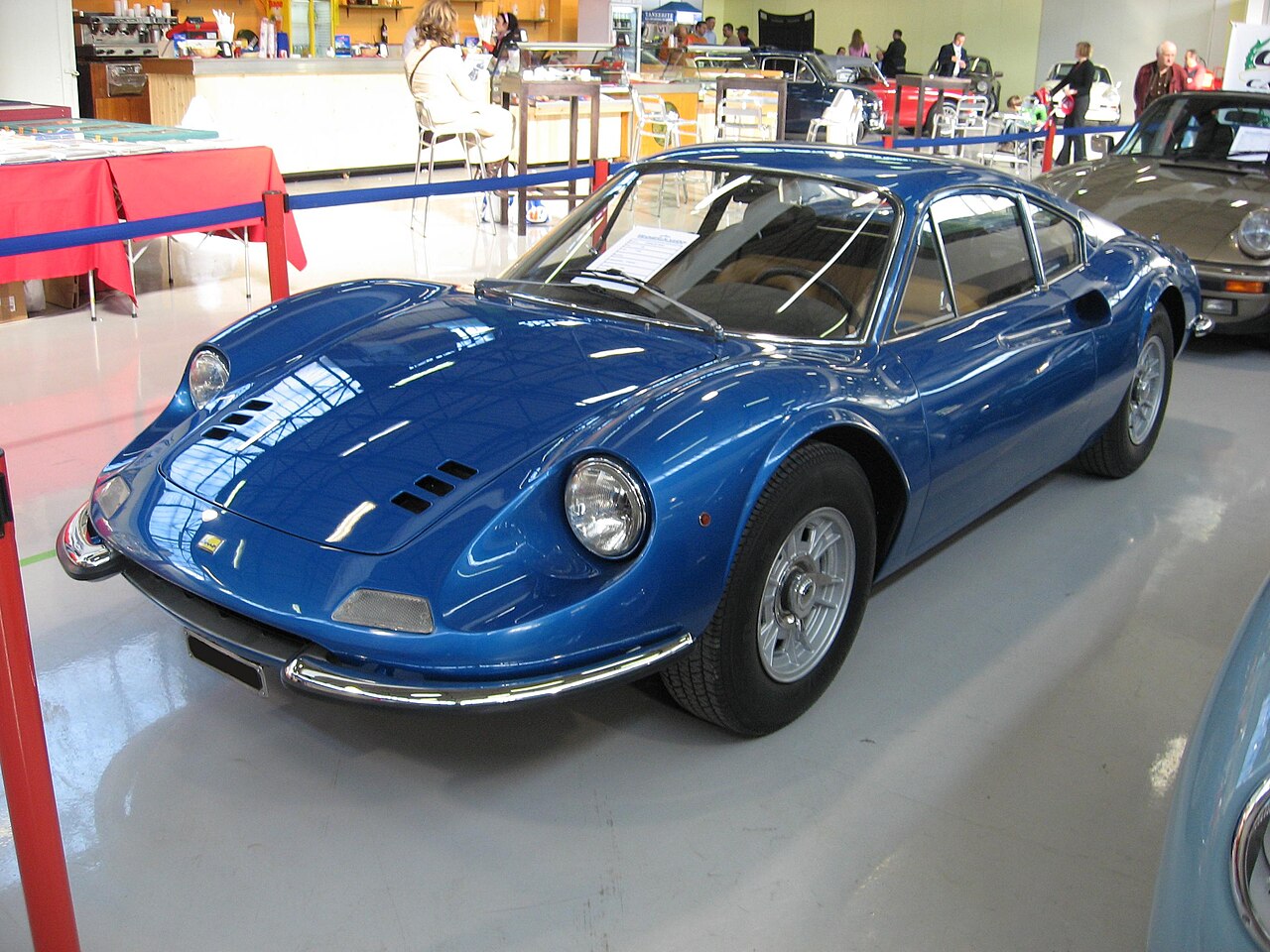 Blue Ferrari Dino 246 GT coupe, front left three-quarter view on the floor of an indoor classic car show.